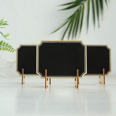 three black and gold frames with wooden legs on a white table next to a plant