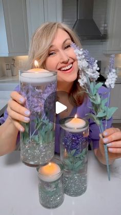 a woman sitting at a table holding two vases filled with flowers and lit candles