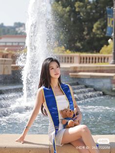 a beautiful young woman sitting next to a fountain holding a teddy bear