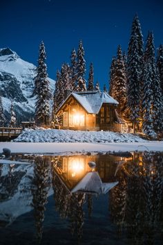 a cabin is lit up at night by the snow covered mountains and lake in front of it