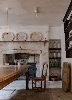 an old fashioned kitchen with stone fireplace and wooden table in the foreground, surrounded by dishes on shelves