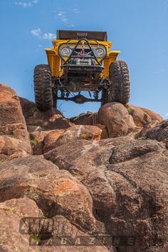 a yellow truck is high in the air over some rocks and boulders with blue skies above