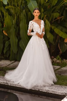 a woman in a white wedding dress standing next to a pool with greenery behind her