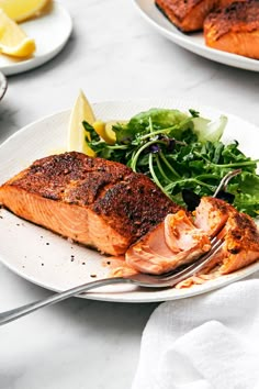 two plates with salmon, greens and lemon wedges on them sitting on a table