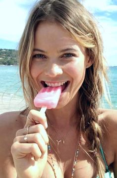 a beautiful young woman holding a candy bar in her hand on the beach with blue sky and water behind her