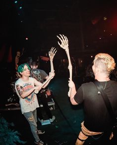 two people with their hands up in the air at a music concert, while one person holds out his hand