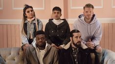 four people sitting on a couch in front of a pink wall and one person standing behind them