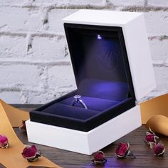 an engagement ring sits in a white box surrounded by rose petals and ribbon on a wooden table