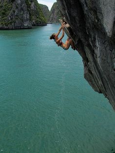 a person climbing up the side of a cliff next to some water and mountains in the background