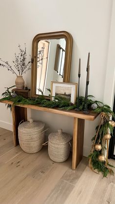 a wooden table topped with lots of greenery next to a mirror and two wicker baskets