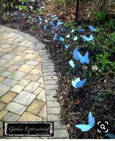 blue paper butterflies are on the ground in front of a brick path and trees with green leaves