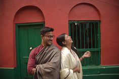 a man and woman standing in front of a red building with green shutters looking up at the sky