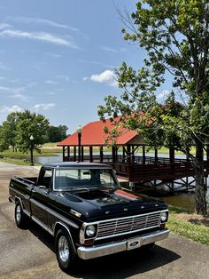 an old black pickup truck parked in front of a lake and wooden bridge with red roof