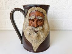 a ceramic mug with a man's face painted on the front and sides, sitting against a white brick wall