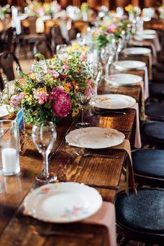 the long table is set with plates and flowers in vases on each side, along with empty wine glasses