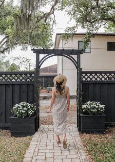 a woman in a dress and hat walking down a brick path through a gated area