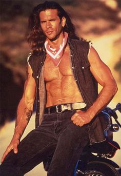 a man with long hair and no shirt on sitting on a motorbike in the desert