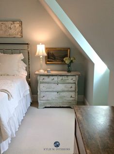 a white bed sitting under a window next to a wooden dresser and lamp in a bedroom