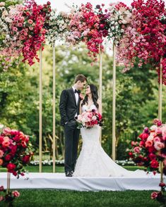 a bride and groom are standing under an arch with pink flowers on the grass in front of them