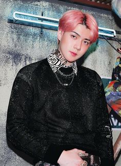 a young man with pink hair wearing a black shirt and silver necklace standing in front of a wall