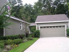 a gray house with trees and bushes around it's front yard, along with a driveway leading to the garage