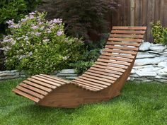 a wooden lounge chair sitting on top of grass next to a rock wall and bushes