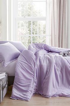 an unmade bed with purple sheets and pillows in front of a large window on a wooden floor