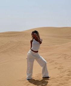 a woman is standing in the sand with her hands behind her head and wearing white pants