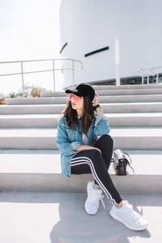 a woman sitting on the steps with her legs crossed and wearing white tennis shoes, jeans jacket and black beanie