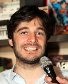 a man holding a microphone in his right hand and smiling at the camera while wearing a blue shirt