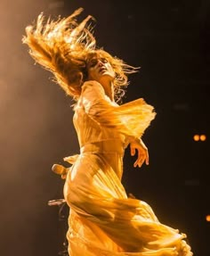 a woman in a yellow dress on stage with her hair blowing in the wind and lights behind her