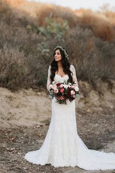 a woman in a wedding dress standing on the side of a dirt road holding flowers