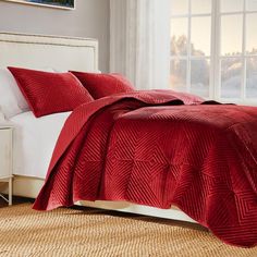 a bed with red bedspread and pillows in a room next to a window