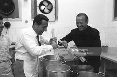 three men in a kitchen preparing food and smiling at the camera while one man is pouring something into a pot