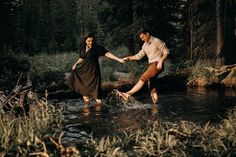 a man and woman crossing a river in the woods with their feet in the water