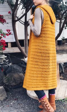 a woman standing in front of a tree while talking on a cell phone and wearing a yellow crocheted vest