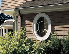 a round window on the side of a brown house with bushes and shrubs around it