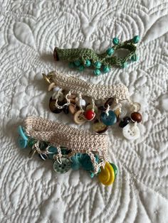 three crocheted bracelets are laying on a white quilted surface with buttons and beads