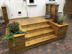 a wooden deck with planters on it next to a white door and brick walkway