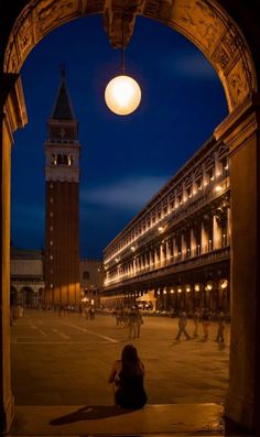 a person sitting on the ground in front of a building at night with a clock tower behind them