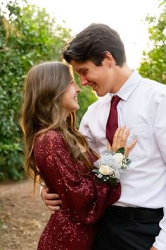 a young man and woman standing next to each other in front of trees with their arms around each other