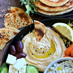 two black plates filled with hummus, olives, cucumbers and carrots