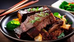 a black plate topped with meat and vegetables next to chopsticks on a wooden table