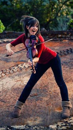 a woman in red shirt playing violin on dirt ground with trees in the backgroud