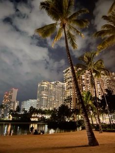 two palm trees on the beach in front of tall buildings at night with clouds overhead