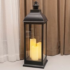 two lit candles in a black lantern on a white table next to a beige curtain