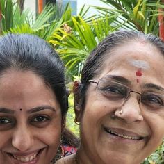 two women are smiling for the camera in front of some plants and trees, one is wearing glasses