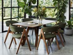 a dining room table surrounded by green chairs and potted plants in front of large windows