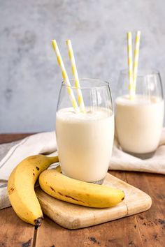 two glasses filled with milk and bananas on top of a wooden table next to each other