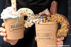 two people holding up cups with ice cream and doughnuts on the top one is filled with sprinkles